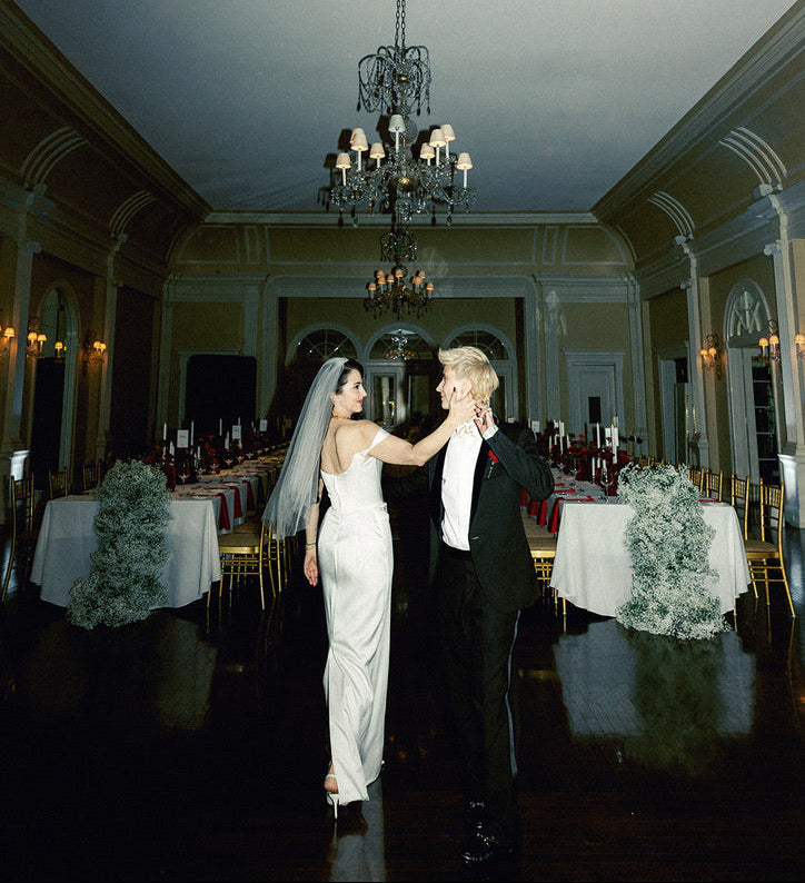 A bride in a white sleeveless gown and veil and a groom in a black tuxedo stand together in a grand ballroom with tables, chandeliers, and warm wall lighting.
